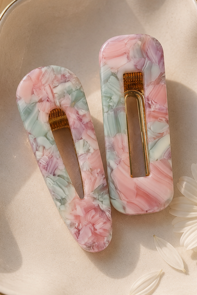 Two rectangular hair claw clips with a pink, green, and white marble pattern, displayed on a scalloped cream ceramic dish with a gold rim, decorated with a white daisy flower and draped with satin fabric.