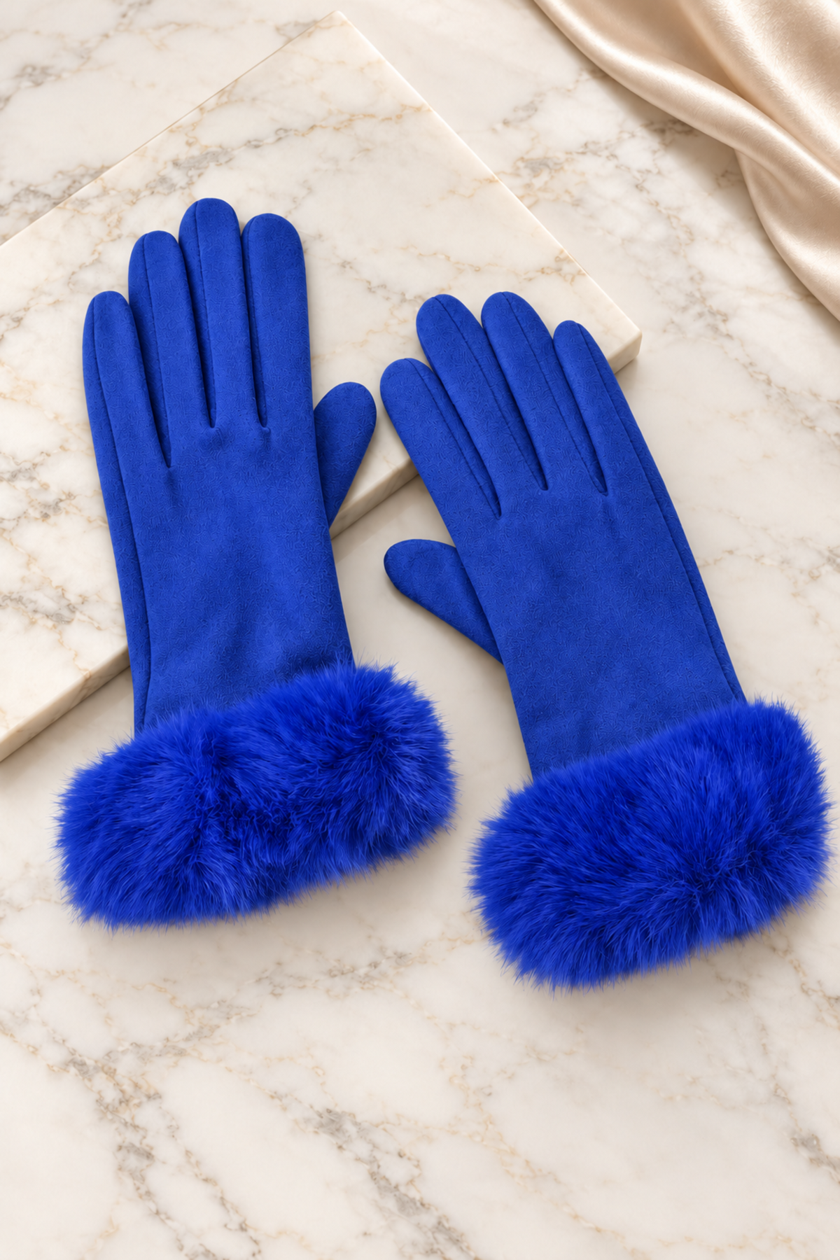 A pair of royal blue gloves with fluffy faux-fur cuffs, displayed on a white marble surface next to a cream-colored book and a draped satin fabric.