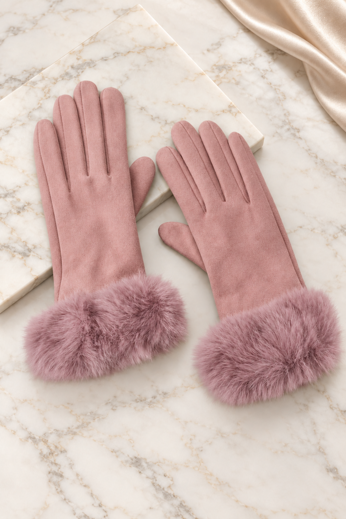 A pair of dusty pink faux-suede gloves with fluffy faux-fur cuffs, displayed on a white marble surface with a cream satin ribbon in the corner.