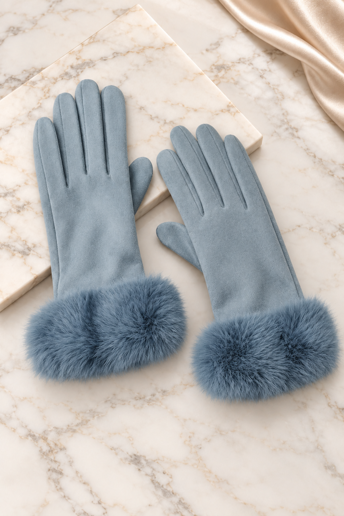 A pair of dusty blue faux-suede gloves with fluffy faux-fur cuffs, displayed on a marble surface next to a cream-colored book and a gold satin ribbon.