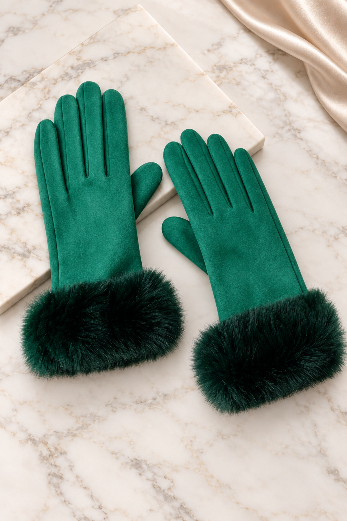 A pair of emerald green gloves with fluffy dark green faux fur cuffs, displayed on a white marble surface next to a cream-colored book and a gold satin ribbon.