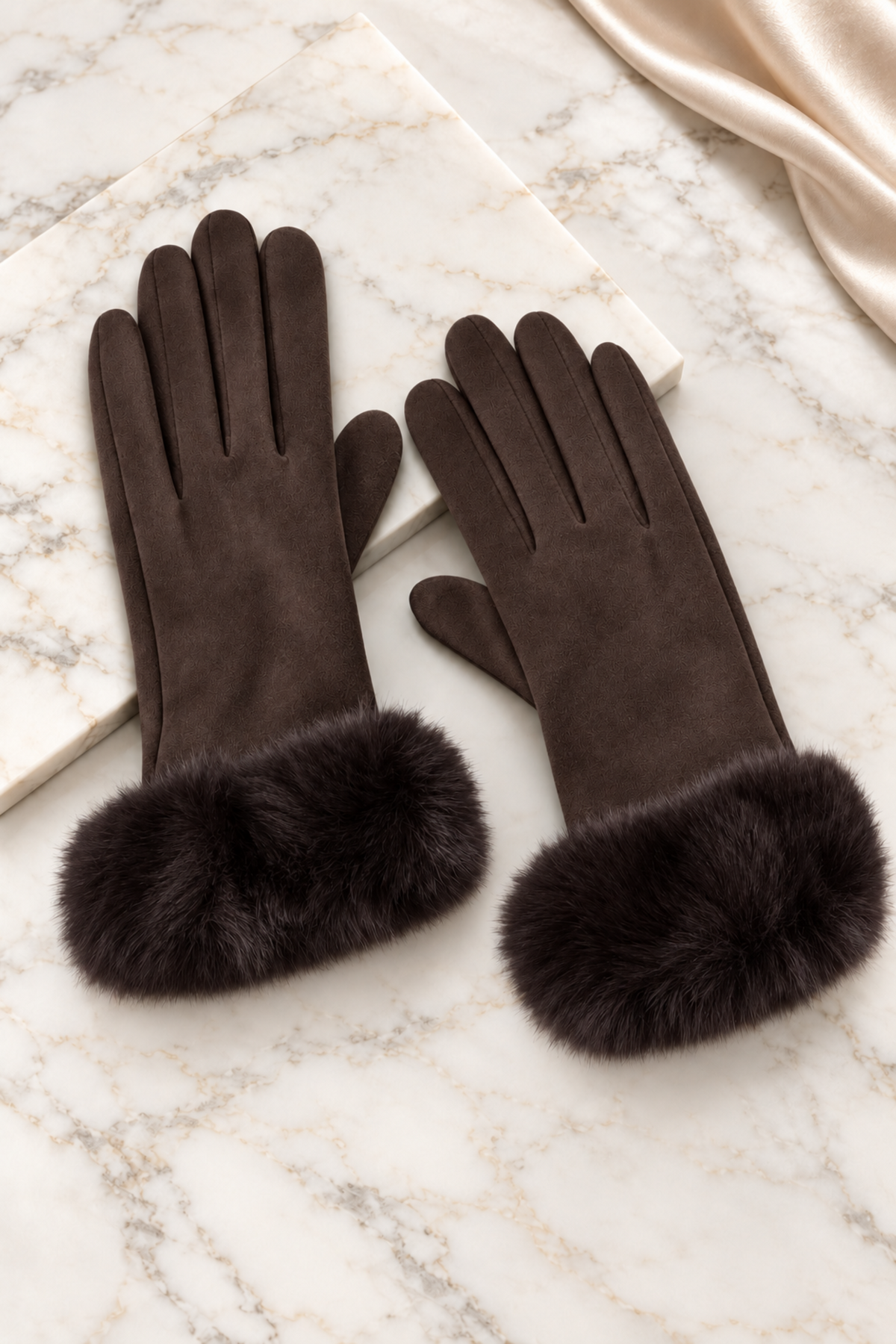A pair of dark brown gloves with plush faux fur cuffs, displayed on a white marble surface with a cream satin fabric in the corner.