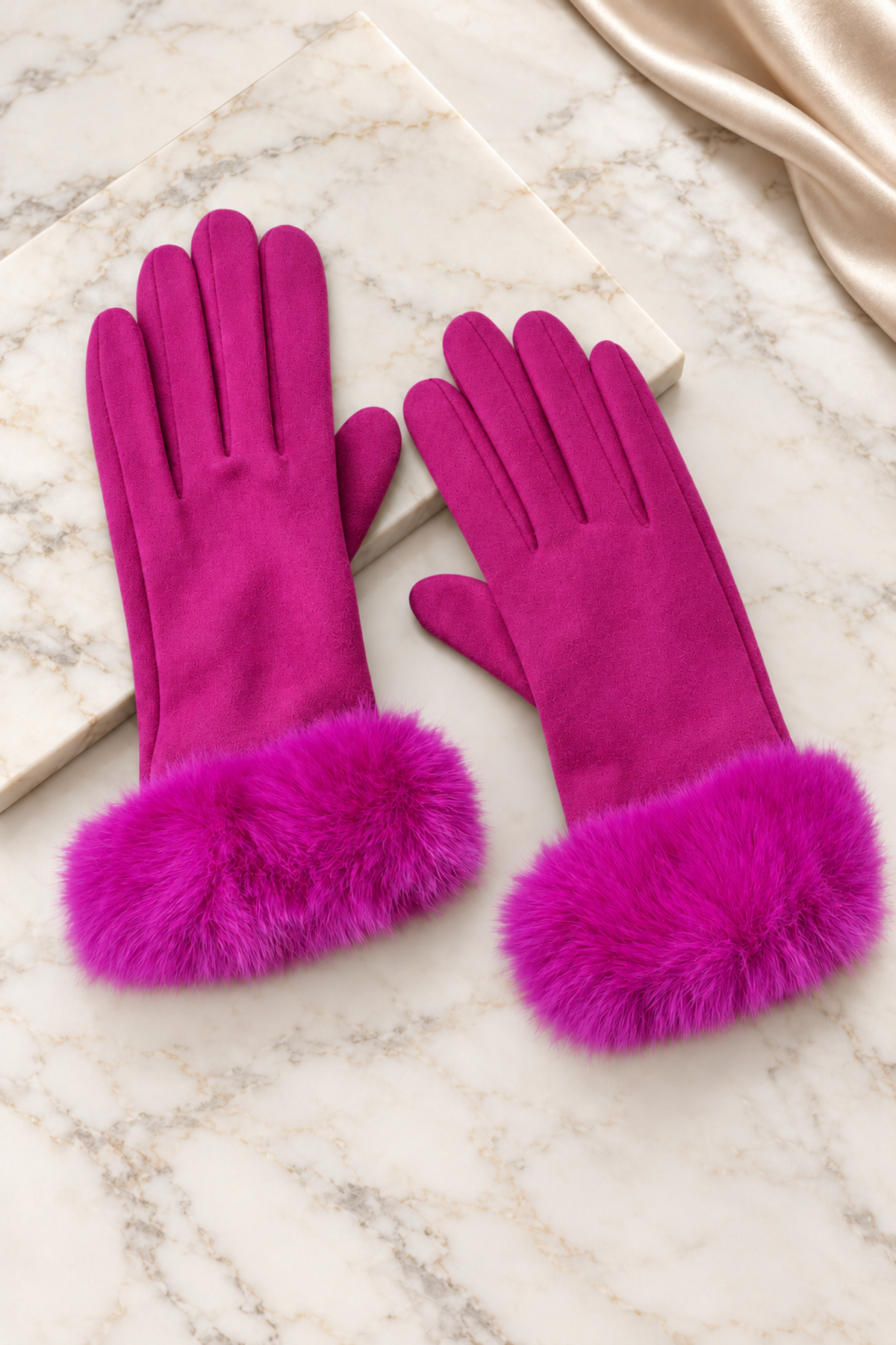 A pair of bright magenta faux-suede gloves with fluffy faux-fur cuffs, displayed on a white marble surface with a cream satin ribbon.