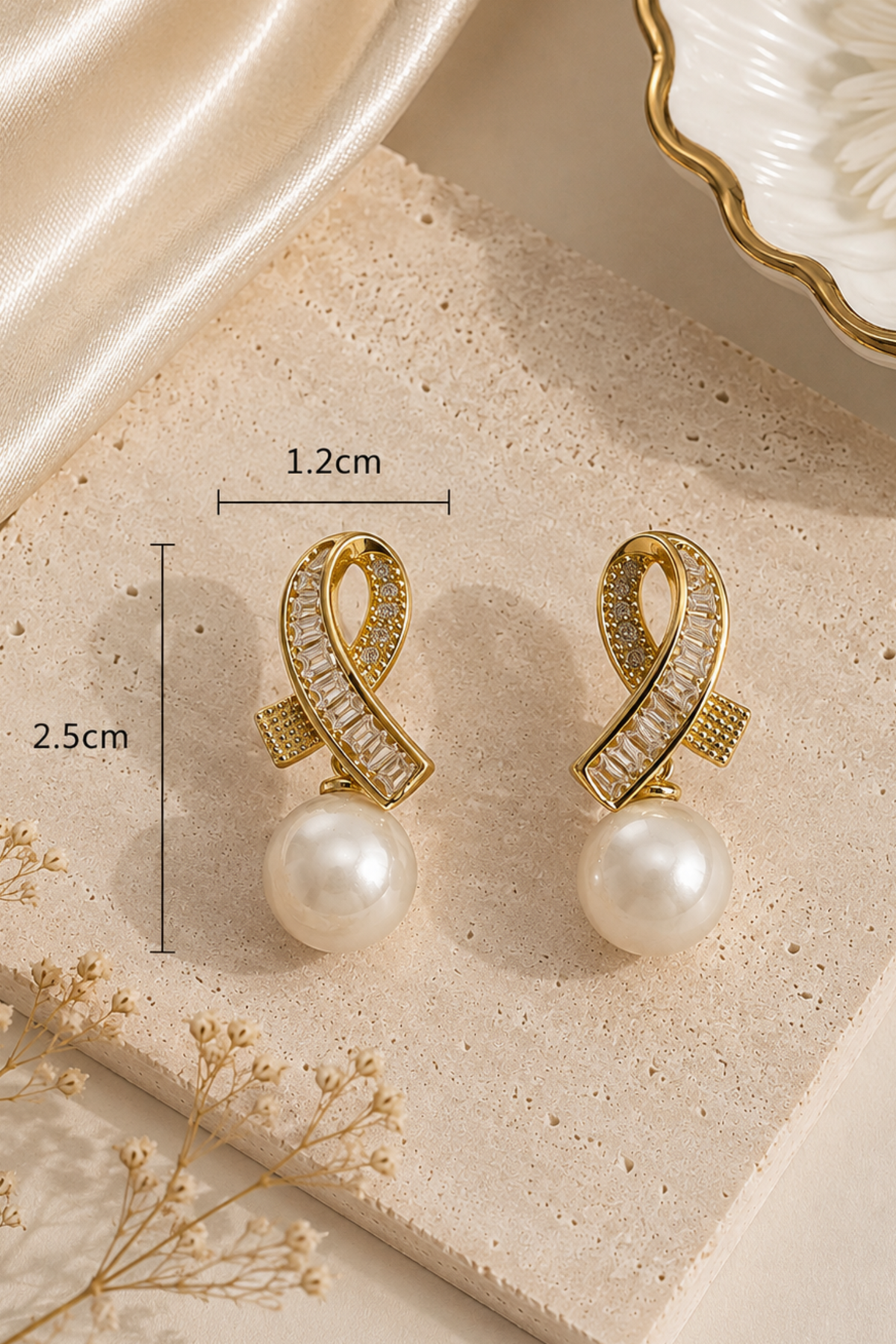 Gold-toned double-loop drop earrings with pavé-set clear stones and a white pearl drop, shown on a beige stone slab with measurement annotations indicating 1.2 cm width and 2.