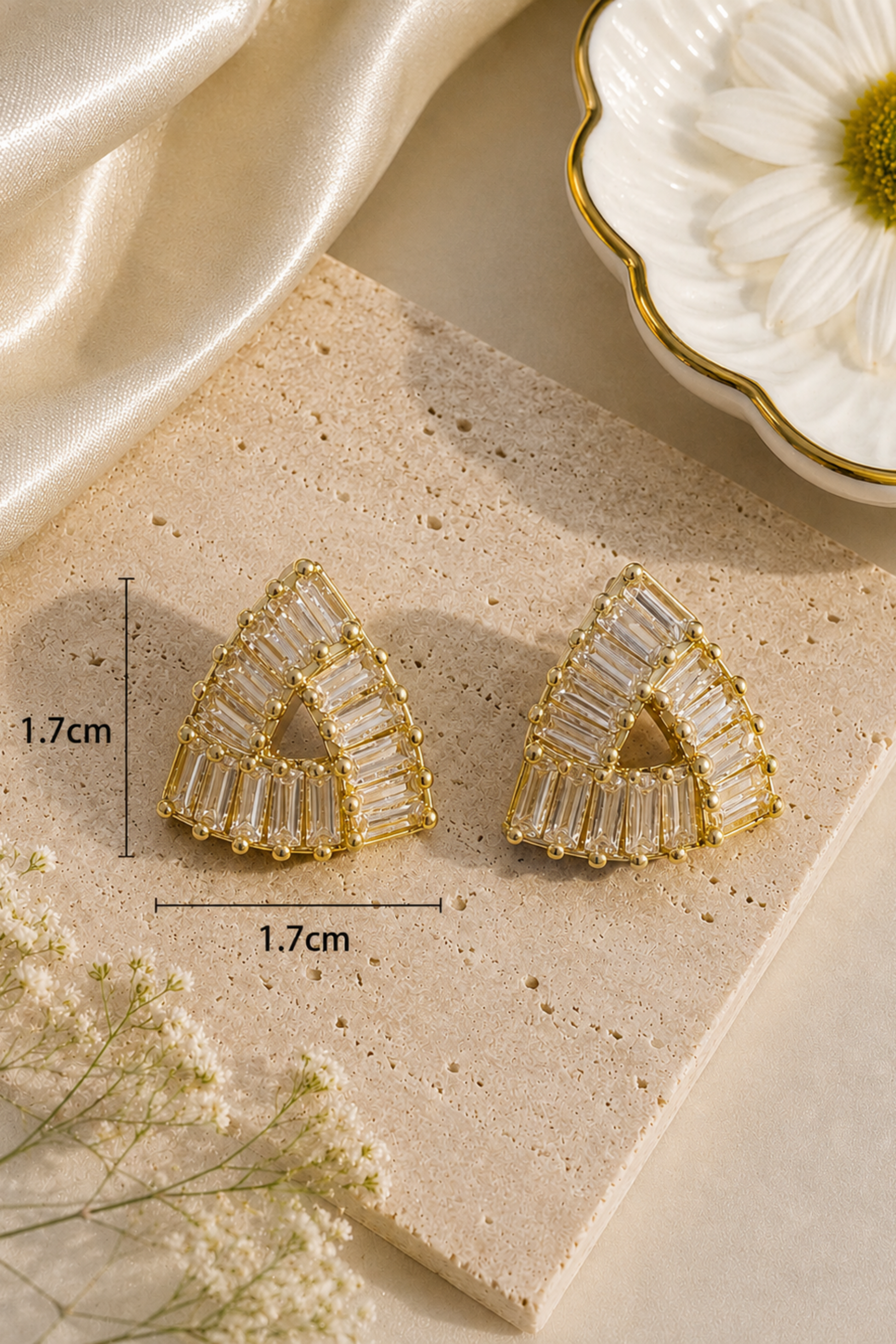 Gold geometric triangle stud earrings with clear rectangular baguette crystals, displayed on a beige stone slab with dimension markers showing 1.7cm by 1.