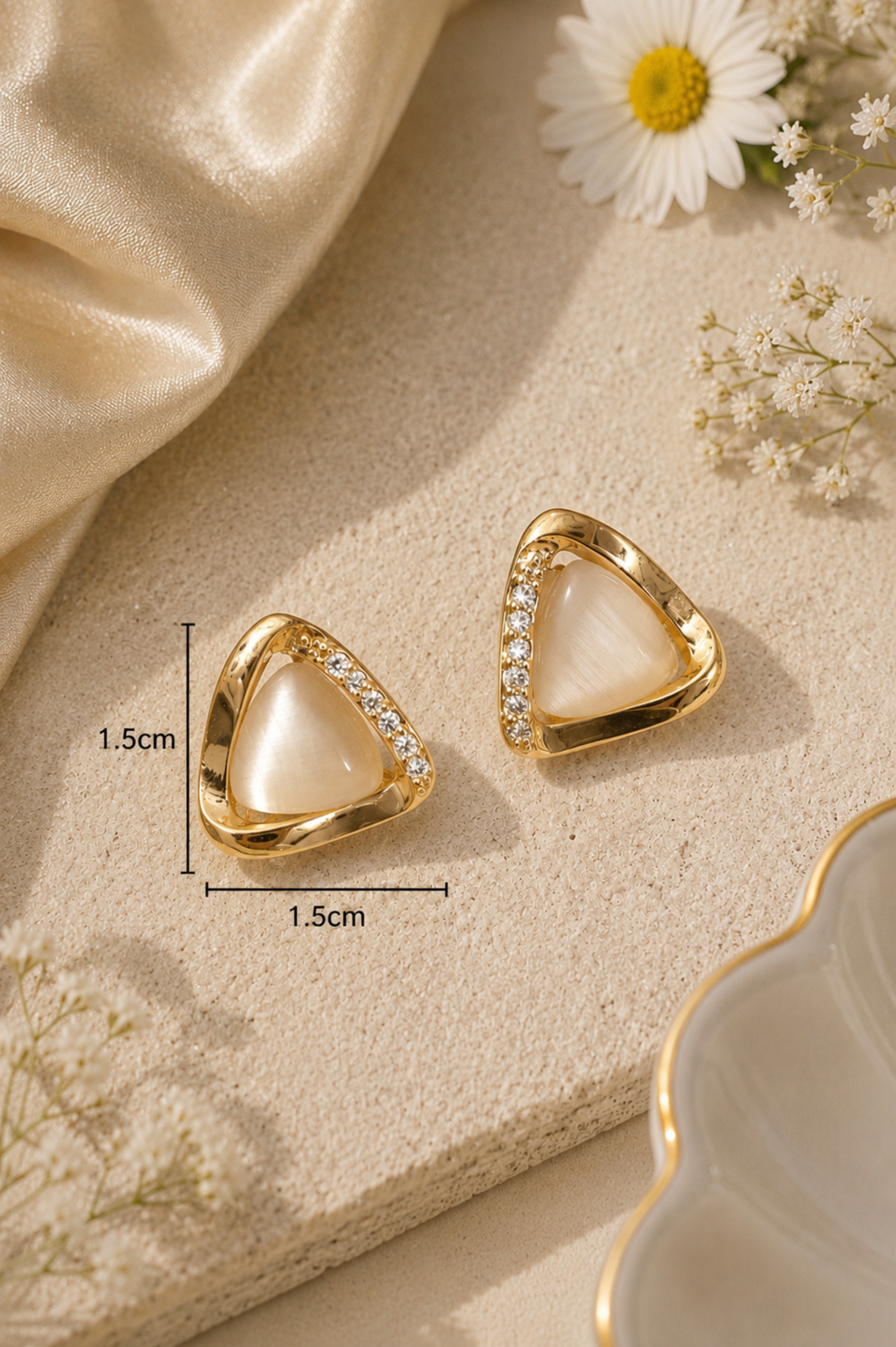 Gold triangular stud earrings with a milky white center and a row of small clear stones, shown on a beige textured surface with decorative flowers and satin fabric. Measurement markers indicate 1.