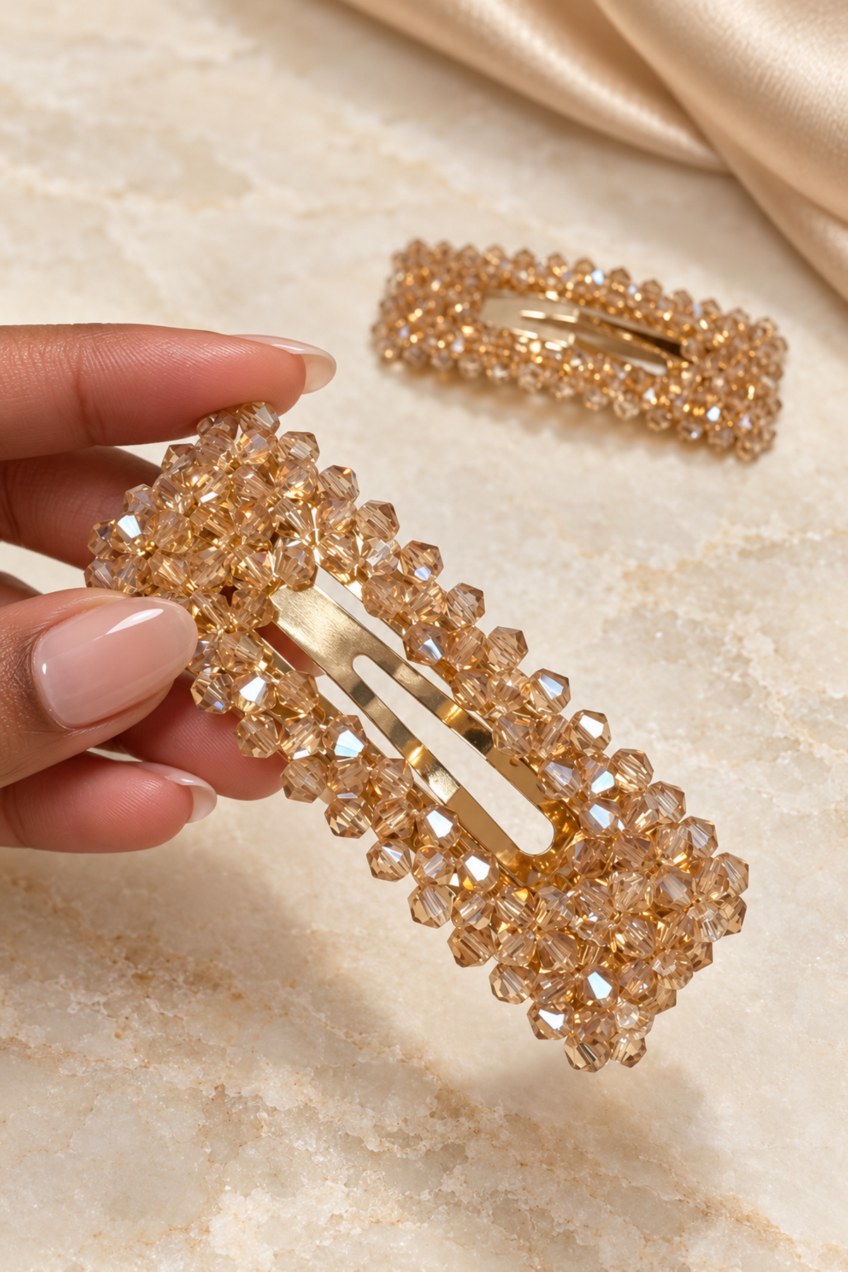 A close-up of a hand holding a gold-tone hair clip embellished with faceted champagne-colored rhinestones, with a second matching clip resting in the background on a beige marble-like surface.