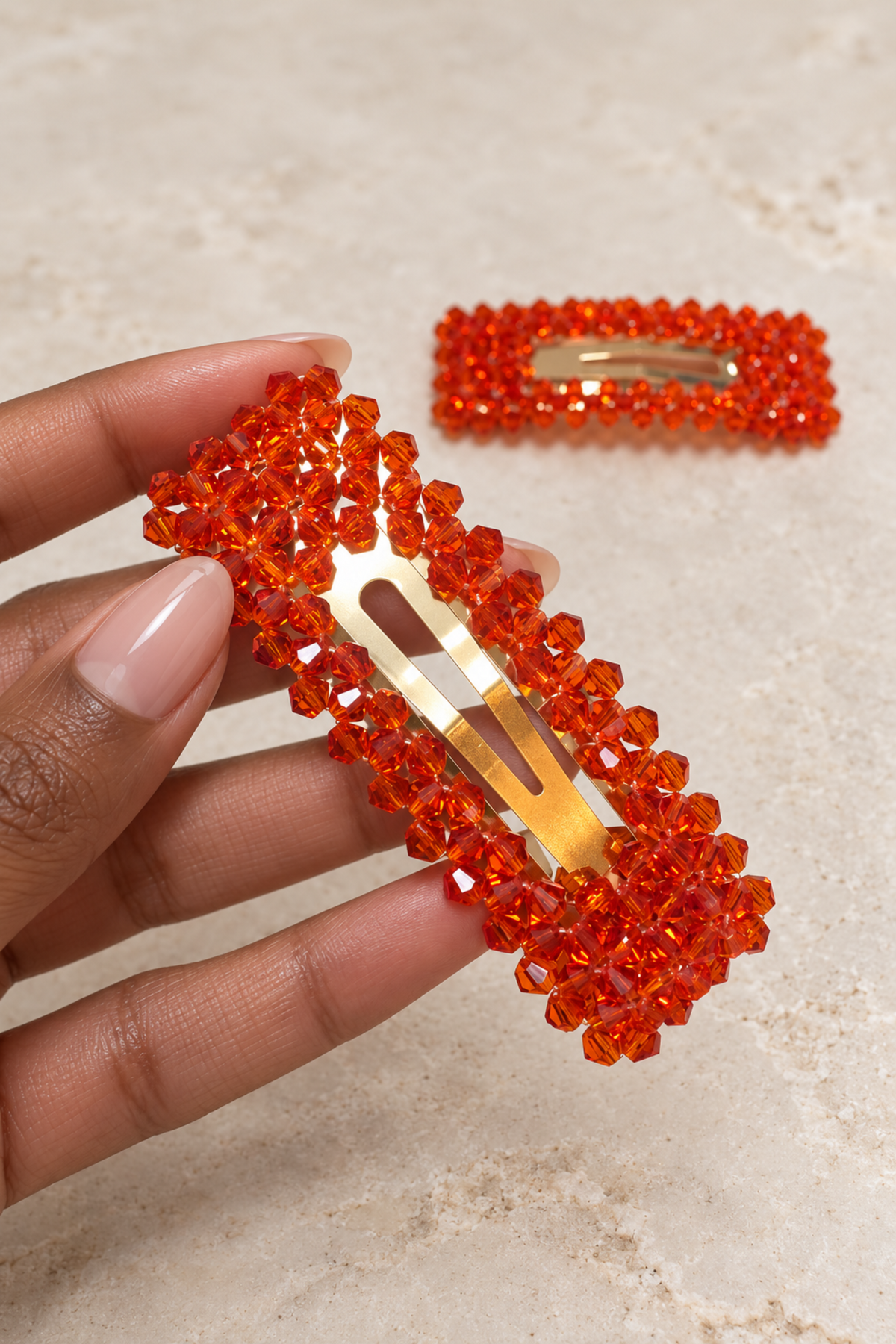 A hand holds a rectangular hair clip covered in bright orange faceted beads, with a second matching clip resting on a light beige stone-like surface in the background.