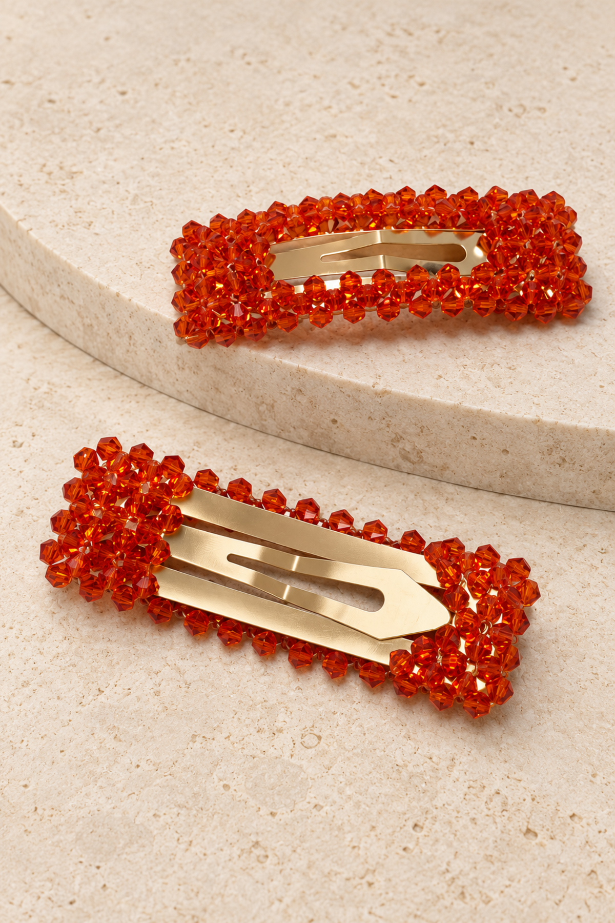Two rectangular hair claw clips embellished with bright orange-red faceted rhinestones and featuring gold-tone metal spring bars, displayed on a light beige textured surface.
