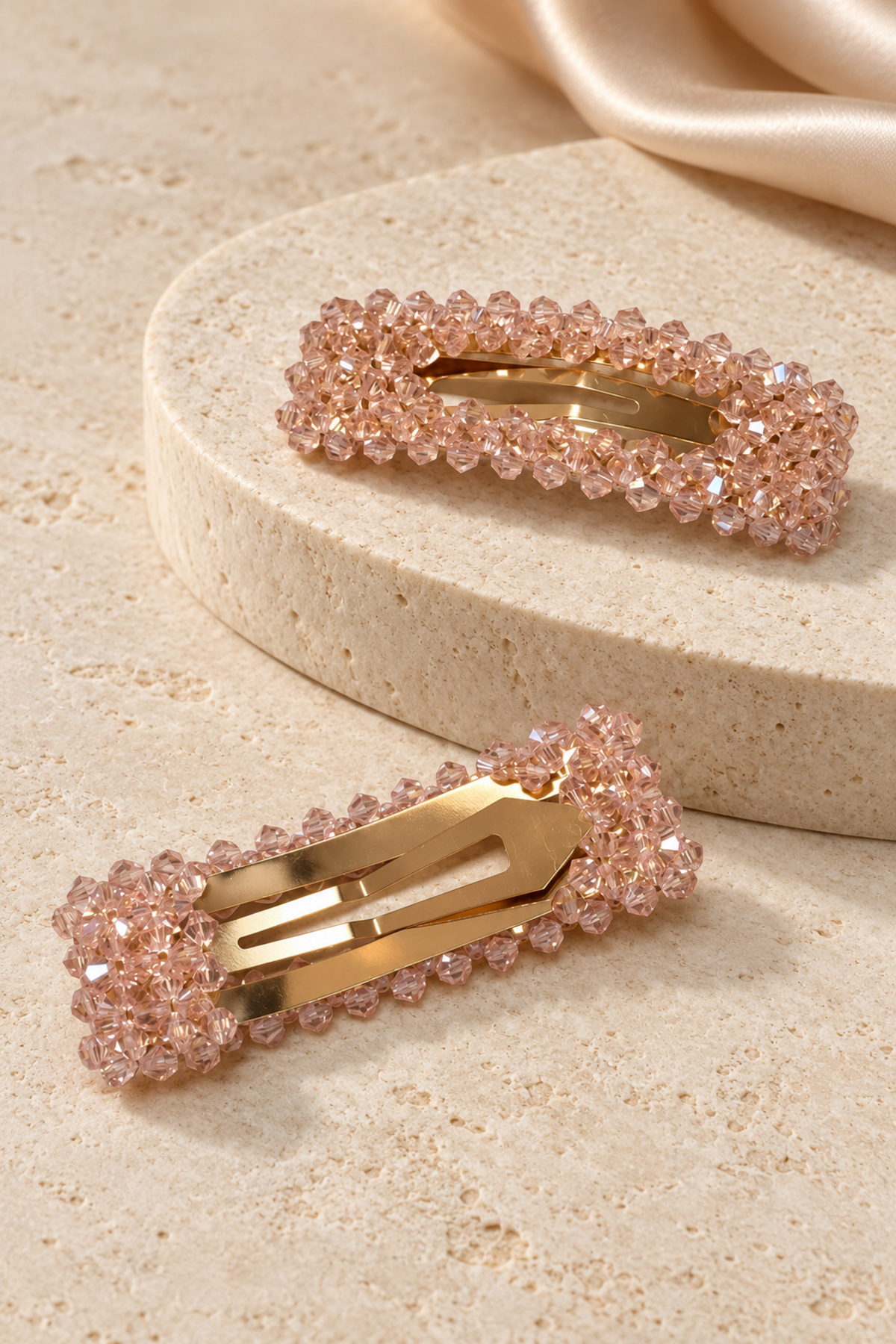 Two rectangular hair claw clips embellished with light pink faceted rhinestones and gold-tone metal, displayed on a beige stone surface with a cream satin fabric in the background.