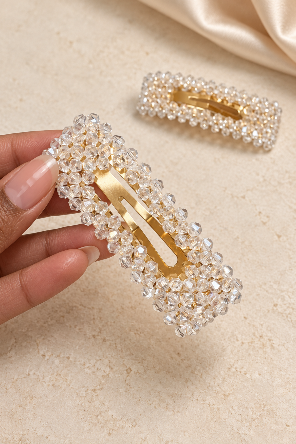 A hand holds a rectangular hair clip adorned with clear rhinestones around a gold-tone metal center, with a second matching clip blurred in the background on a light beige textured surface.