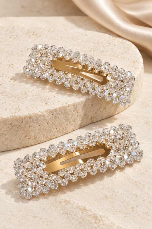 Two rectangular hair claw clips embellished with clear rhinestones, showcasing a gold-tone metal interior, arranged on a textured beige stone surface with a draped satin fabric in the background.
