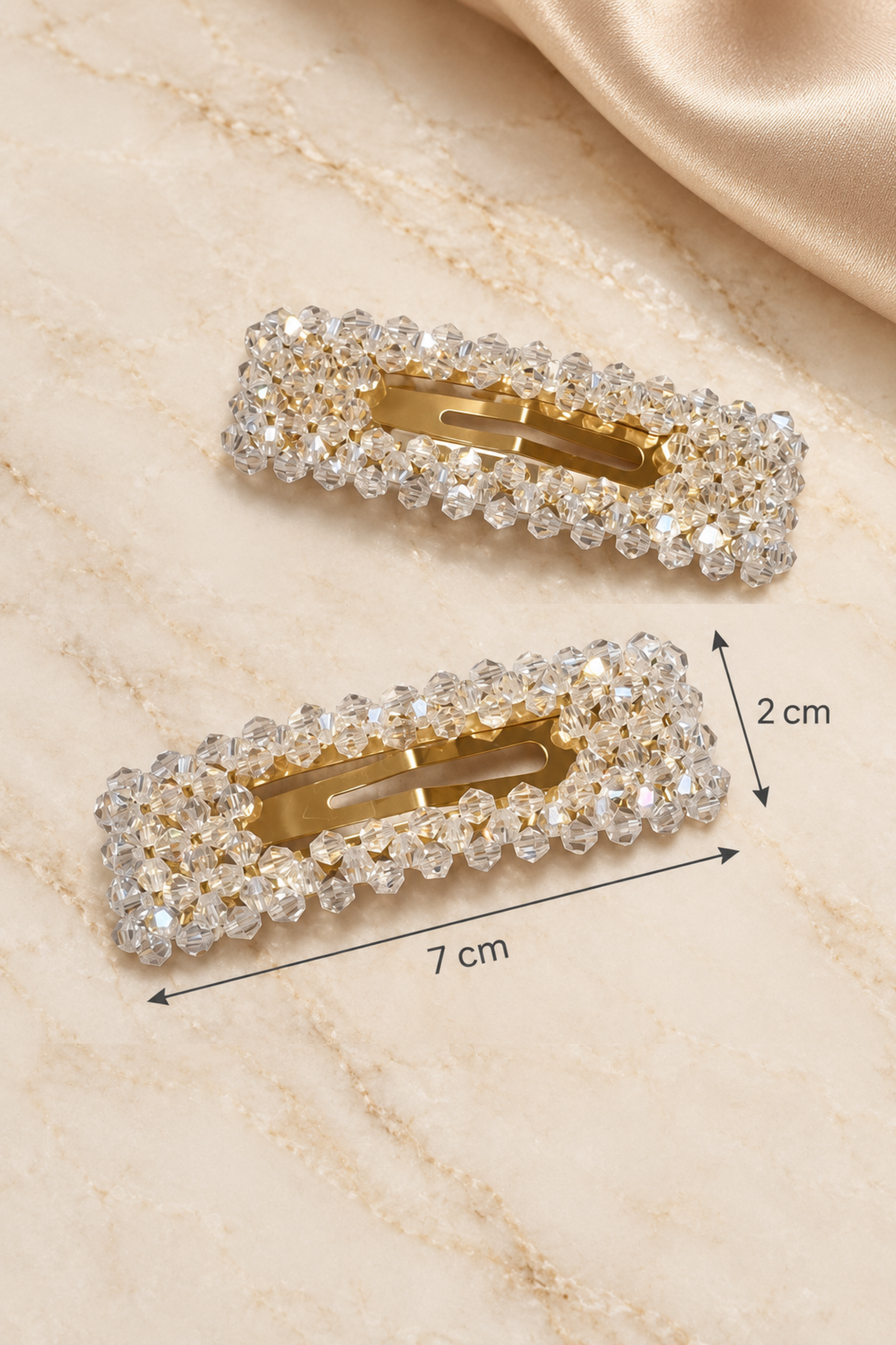 Two rectangular hair barrettes covered in clear rhinestones with gold-tone metal interiors, shown on a light beige marble surface with a satin fabric corner. Black arrows and labels indicate dimensions: 7 cm length and 2 cm width.