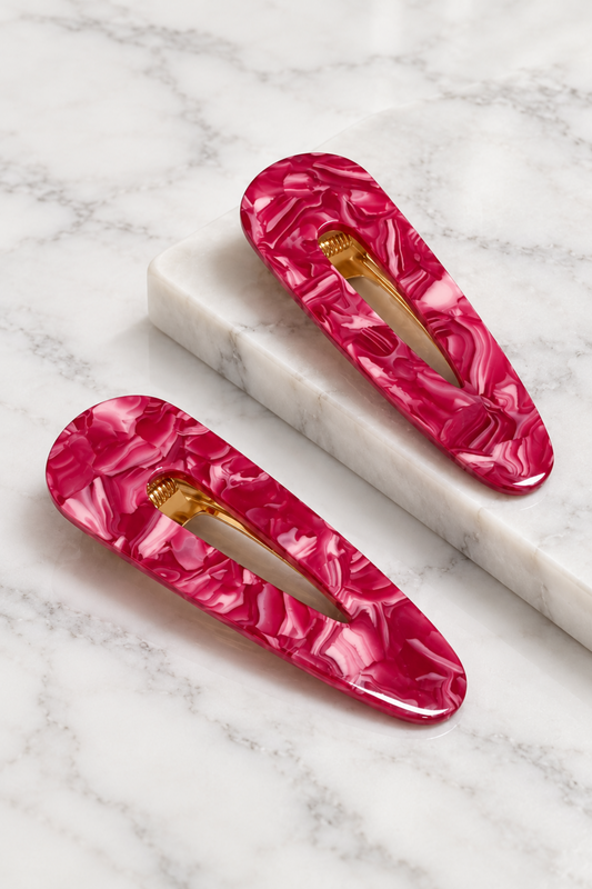 Two glossy hot pink marbled hair claw clips with gold metal spring bars, displayed on a white marble block over a white marble surface.