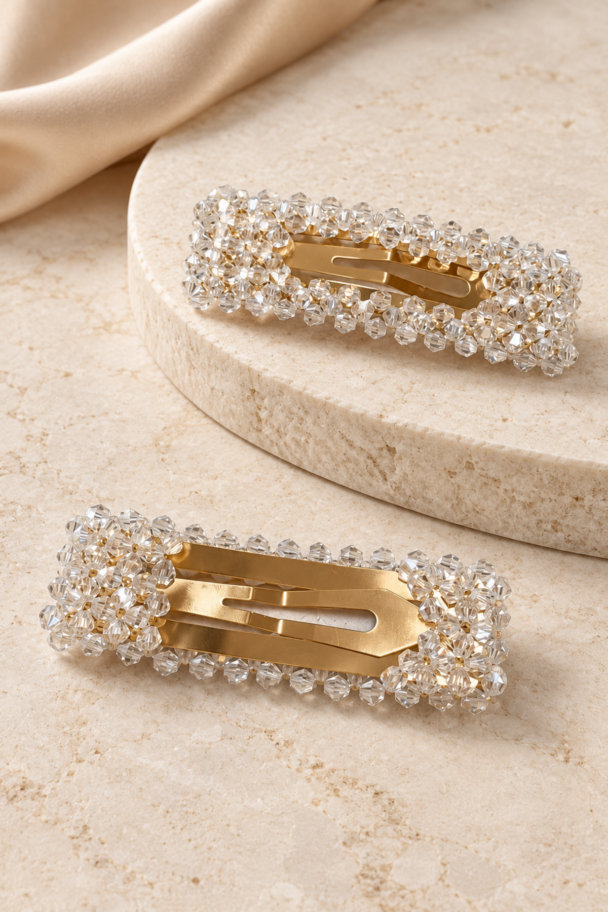 Two gold hair claw clips embellished with clear rhinestones, displayed on a beige stone surface with a draped beige fabric in the background.