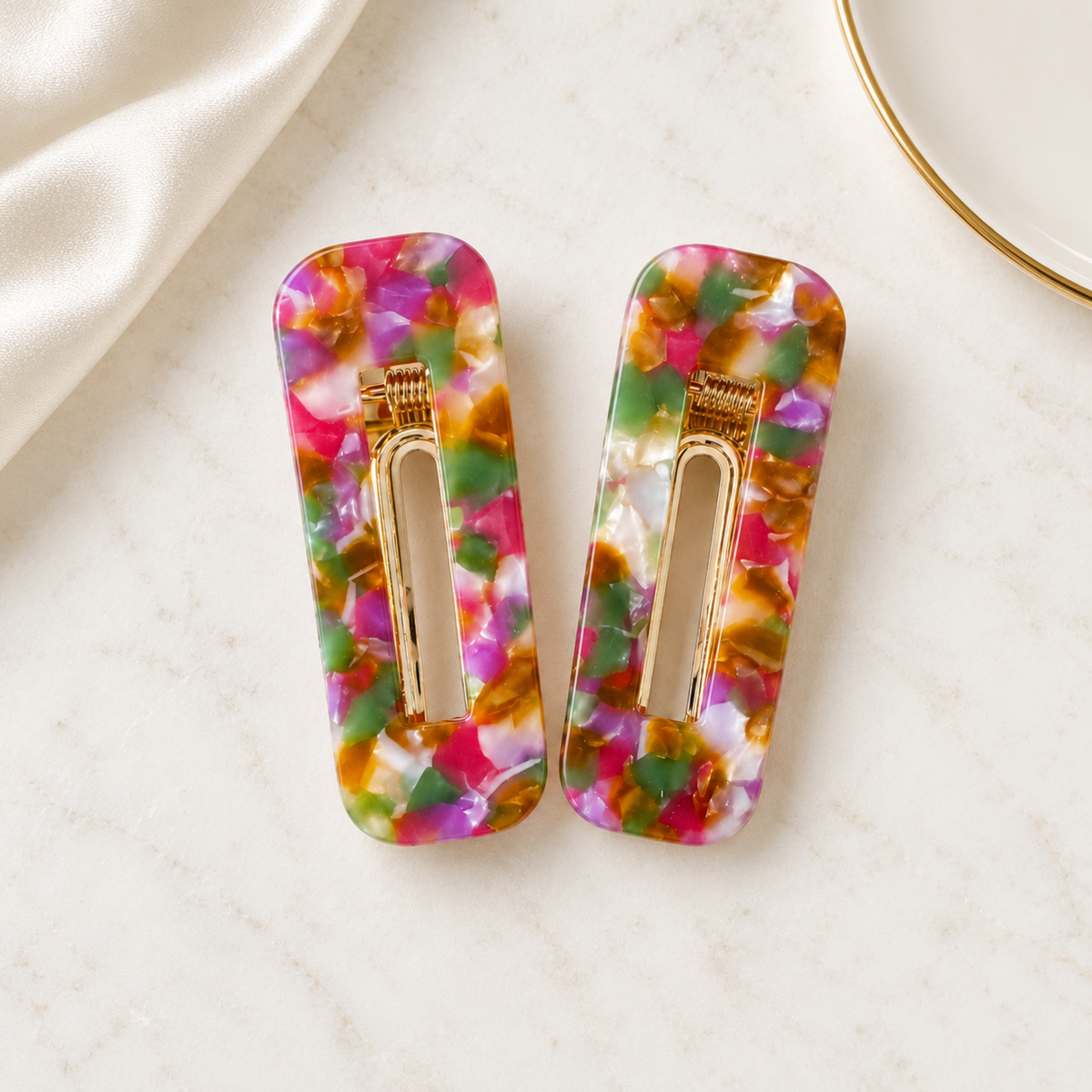 A pair of rectangular hair clips with a colorful marbled pattern in pink, green, orange, and white, featuring gold-tone metal spring clips, displayed on a light marble surface with a white satin cloth and a gold-rimmed plate.