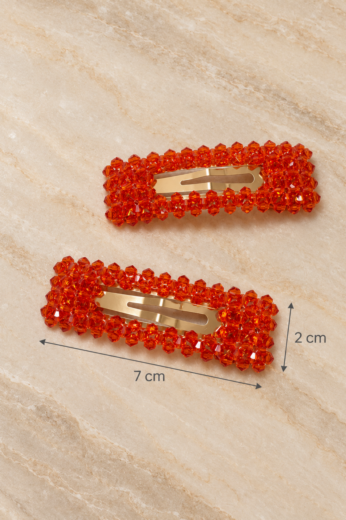 Two rectangular hair clips covered in bright orange-red rhinestones with gold-tone metal clasps, shown on a light beige marble-like surface with dimension labels indicating 7 cm length and 2 cm height.