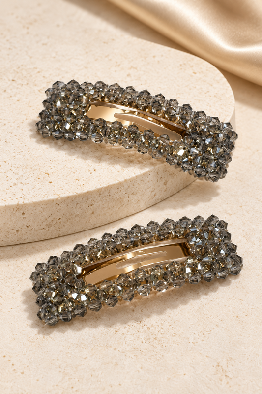 Two rectangular barrette hair clips with clear faceted rhinestones and a gold-tone center, displayed on a beige stone pedestal with a gold satin fabric in the background.