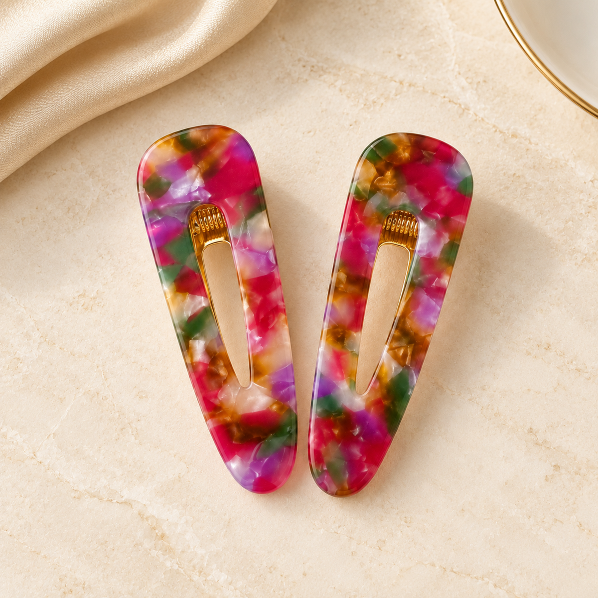 Two rectangular hair clips with a vibrant, multicolored tie-dye pattern in pink, green, purple, and gold, displayed on a beige textured surface with a cream satin fabric in the corner.