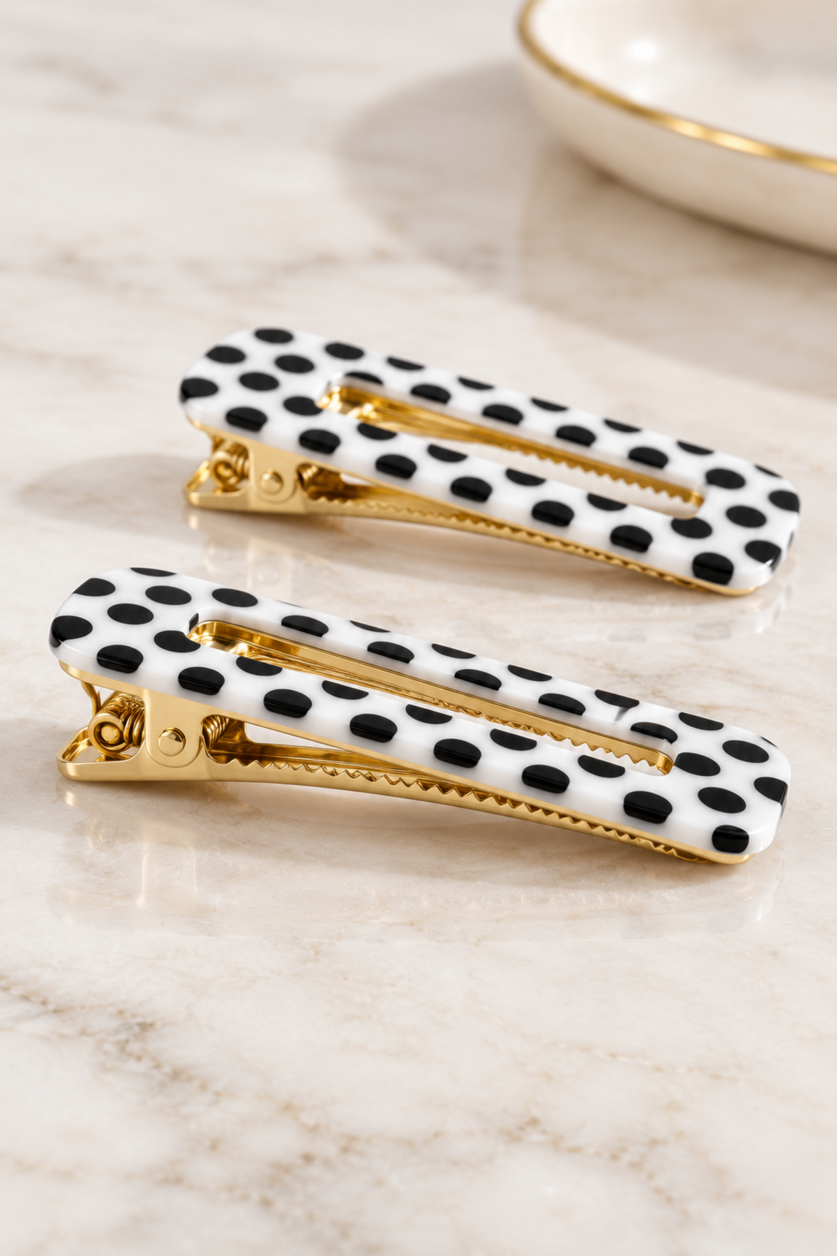 Two black and white polka dot hair clips with gold-tone metal clasps, displayed on a light marble surface.