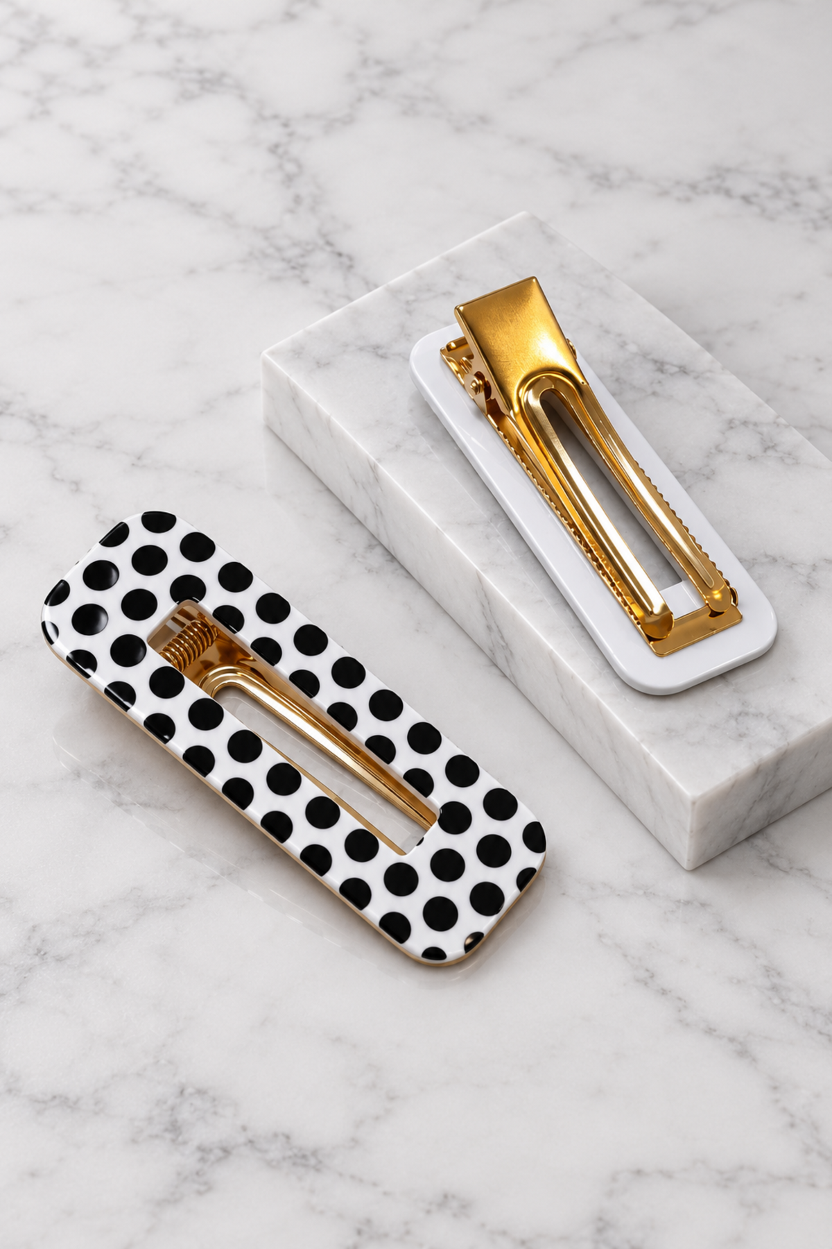 Two gold-tone hair claw clips displayed on a white marble surface, one with a white base and black polka dots and one with a white base.