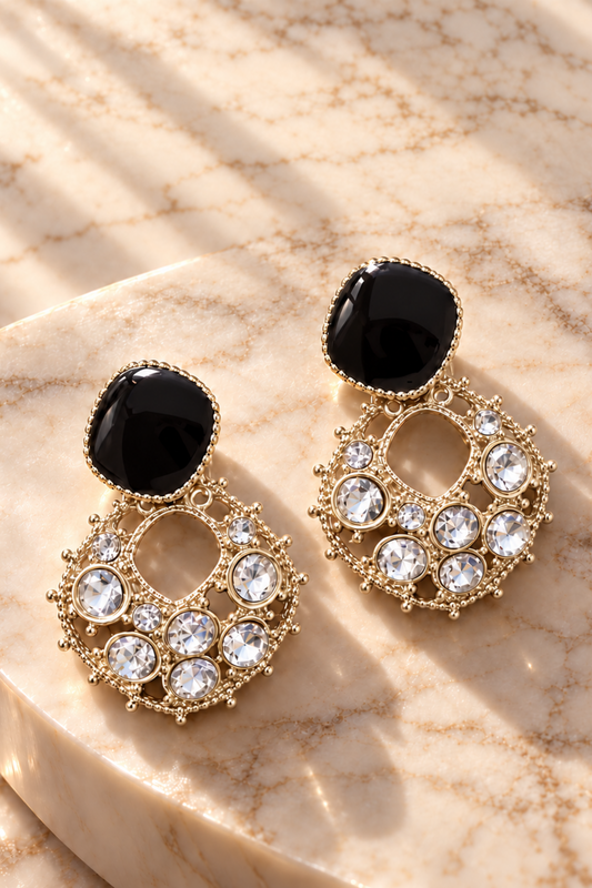 A pair of gold-tone dangle earrings with black oval stones and clear rhinestone clusters, displayed on a beige marble slab with soft shadows.