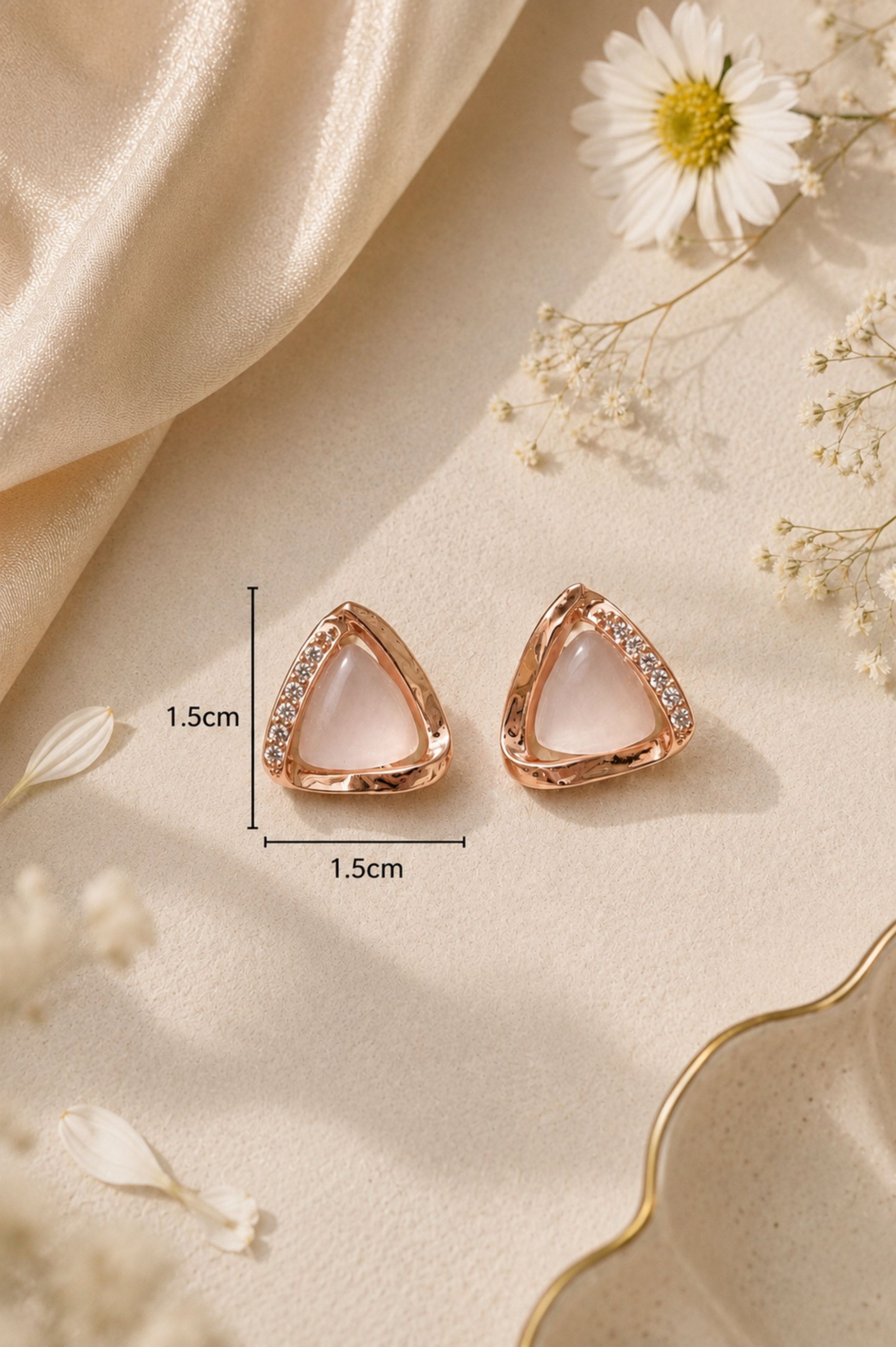 Rose gold triangular stud earrings with a clear crystal border and a pale pink center, shown on a beige fabric background with decorative dried flowers and dimension lines indicating 1.5cm by 1.