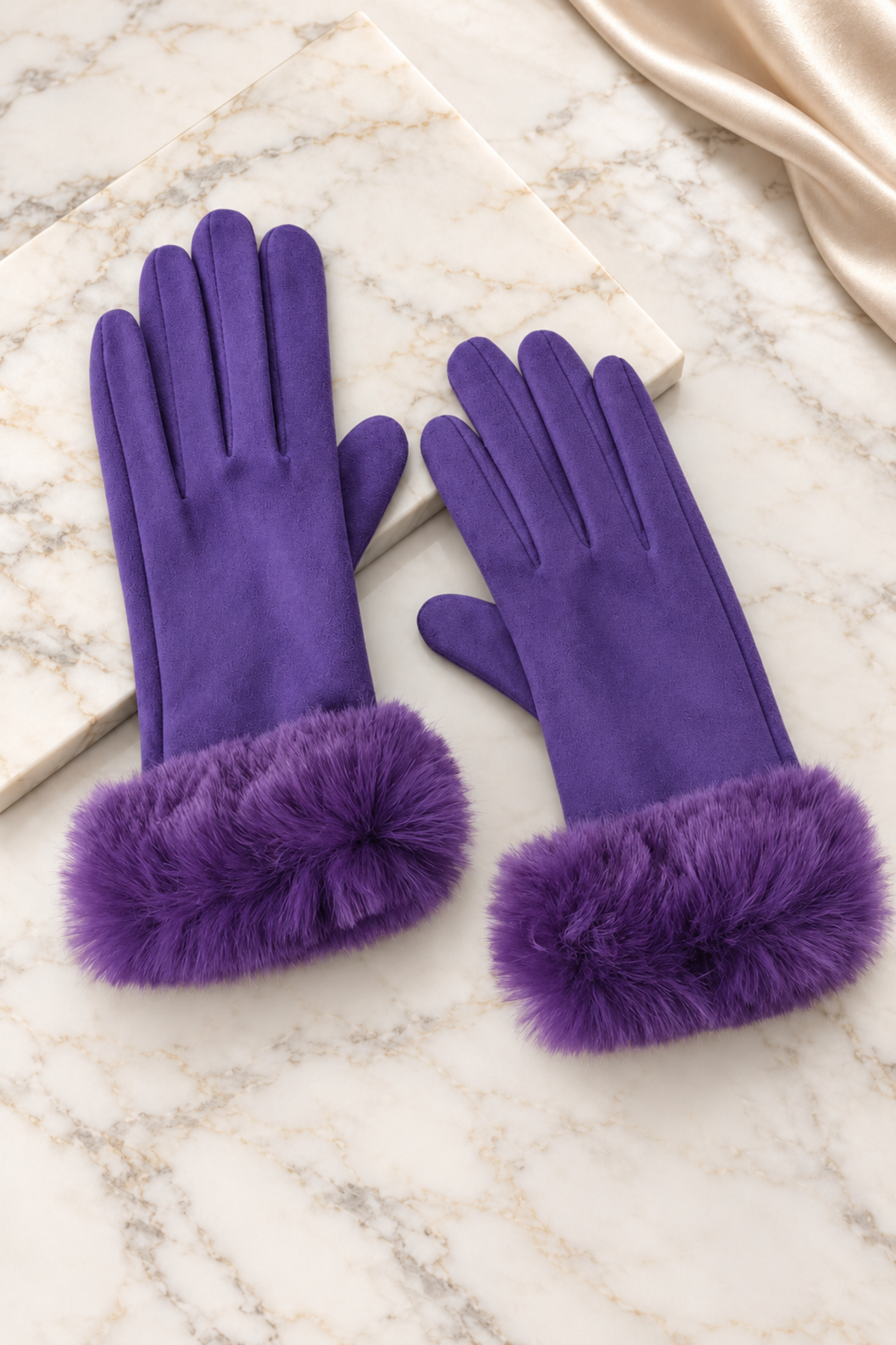 A pair of purple gloves with fluffy faux-fur trim at the cuffs, arranged on a light marble surface with a cream satin fabric in the corner.