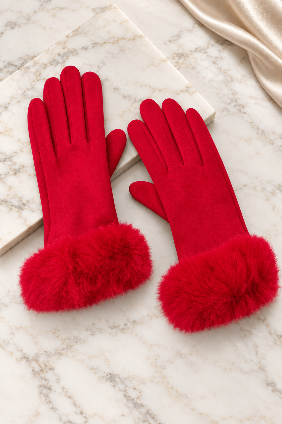 A pair of bright red gloves with fluffy faux-fur cuffs, displayed on a white marble surface next to a cream satin fabric.