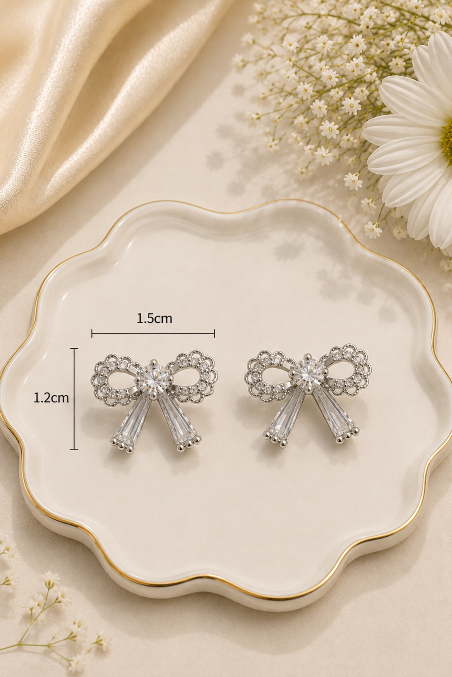 Silver bow-shaped stud earrings with clear rhinestone accents and clear crystal drops, displayed on a scalloped cream dish with dimension markers showing 1.5cm width and 1.