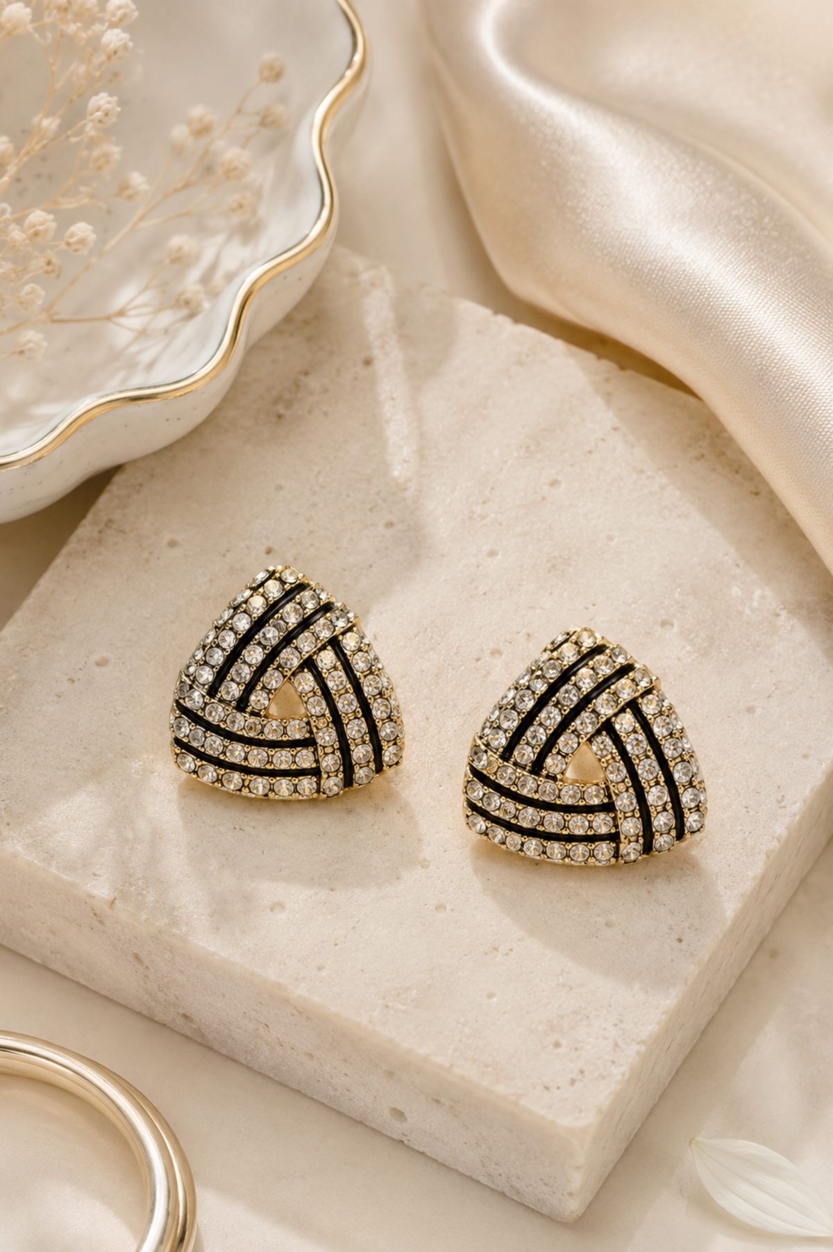 Triangular stud earrings with black and gold striped patterns and clear crystal accents, displayed on a cream stone block with gold-toned jewelry props.