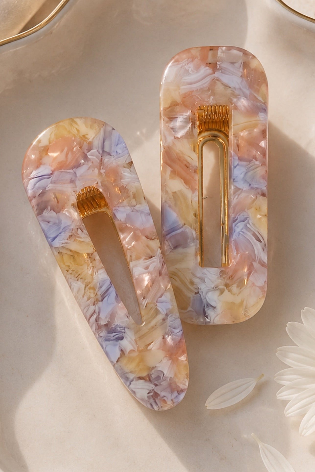 Two acrylic hair claw clips with a marbled floral pattern in pastel orange, yellow, and lavender, displayed on a scalloped cream ceramic dish with a gold rim alongside a white daisy flower.