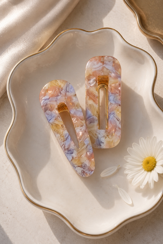 Two acrylic hair claw clips with a marbled floral pattern in pastel orange, yellow, and lavender, displayed on a scalloped cream ceramic dish with a gold rim alongside a white daisy flower.