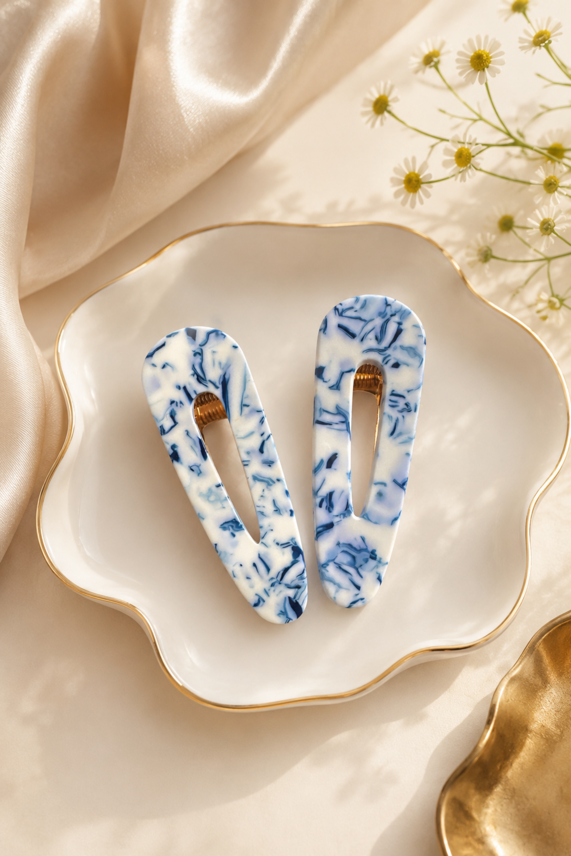 Two blue and white marbled hair clips displayed on a scalloped white ceramic dish with gold trim, styled with beige satin fabric and small daisy flowers.