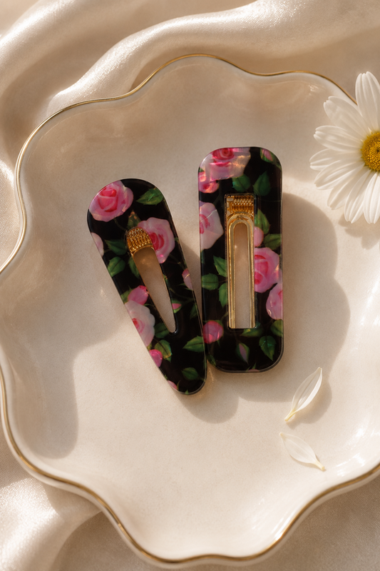 Two floral hair claw clips with pink rose patterns on a black background, displayed on a scalloped cream ceramic dish with a gold rim, styled with a white daisy flower.