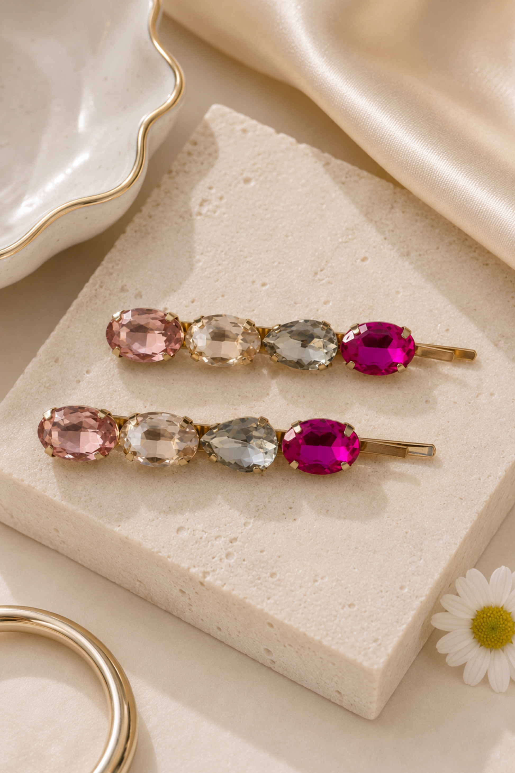 Two gold-toned hair claw clips with pastel and bright gemstones, displayed on a textured cream stone block with elegant fabric and jewelry props.