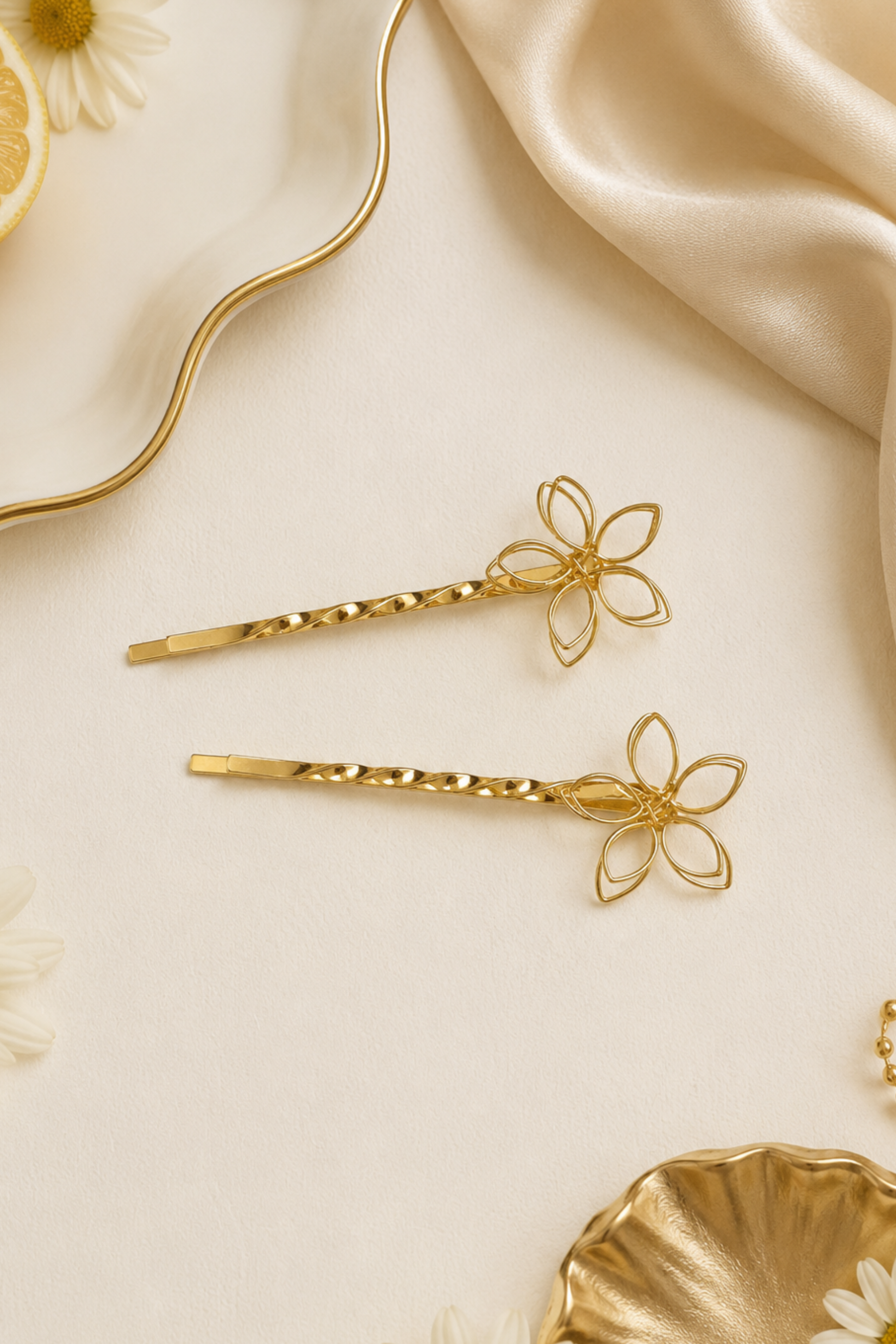 Two gold-tone flower-shaped hair pins arranged on a cream-colored surface with decorative props including white daisies, a lemon slice, draped satin fabric, and a gold dish.