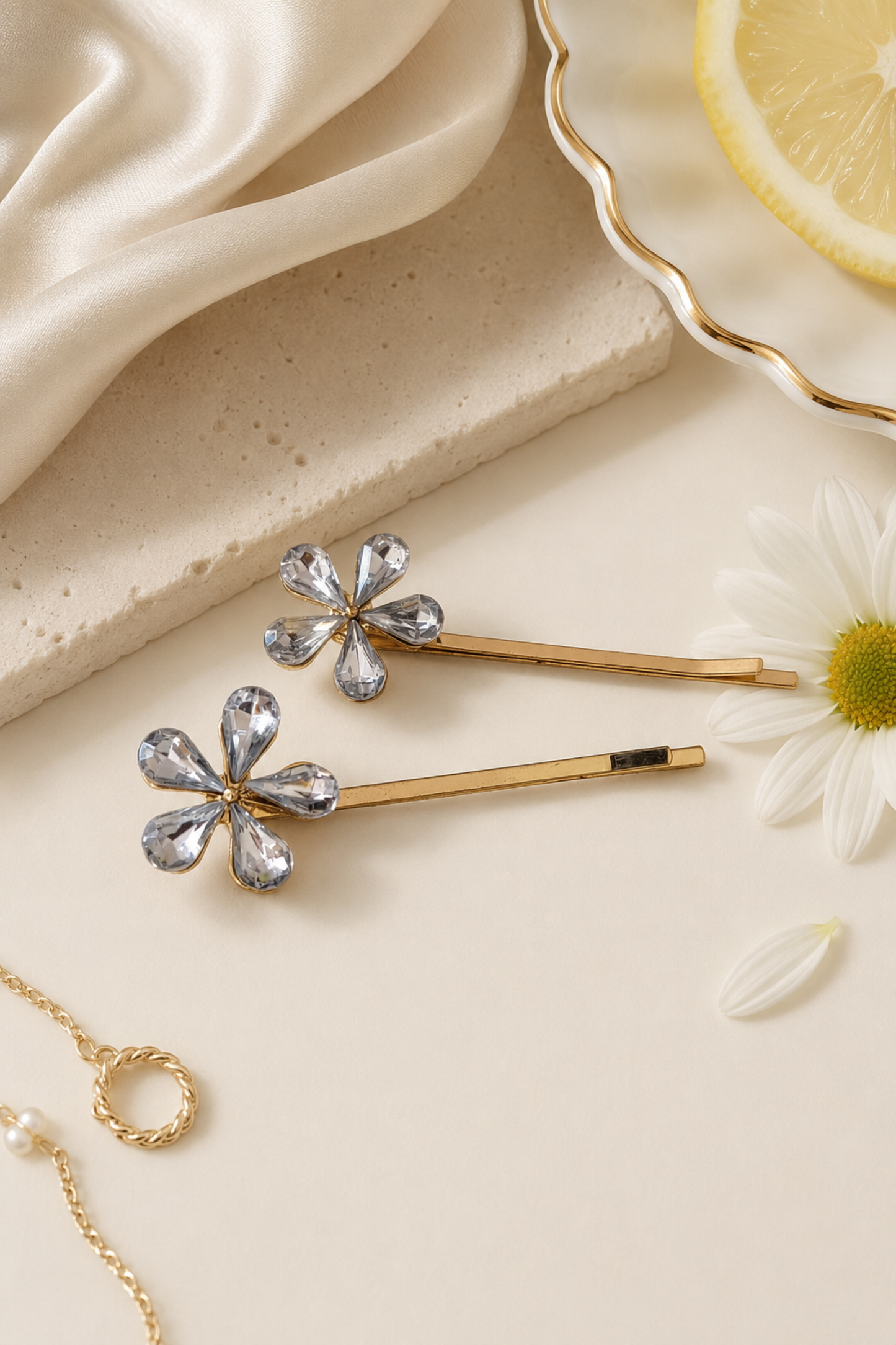 Two gold-tone flower-shaped hair pins with clear rhinestone petals, arranged on a cream surface with a cream daisy, a lemon slice, draped satin fabric, and a gold chain as styled props.