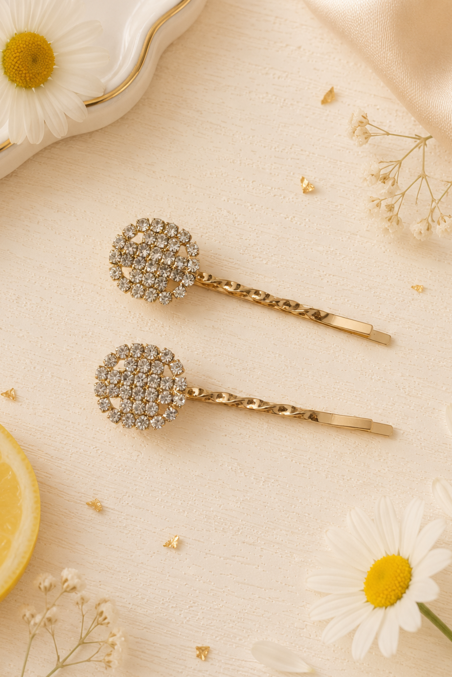 Two gold-tone hair claw clips embellished with round clusters of clear rhinestones, styled on a cream fabric background with daisies, a lemon slice, and a white dish.