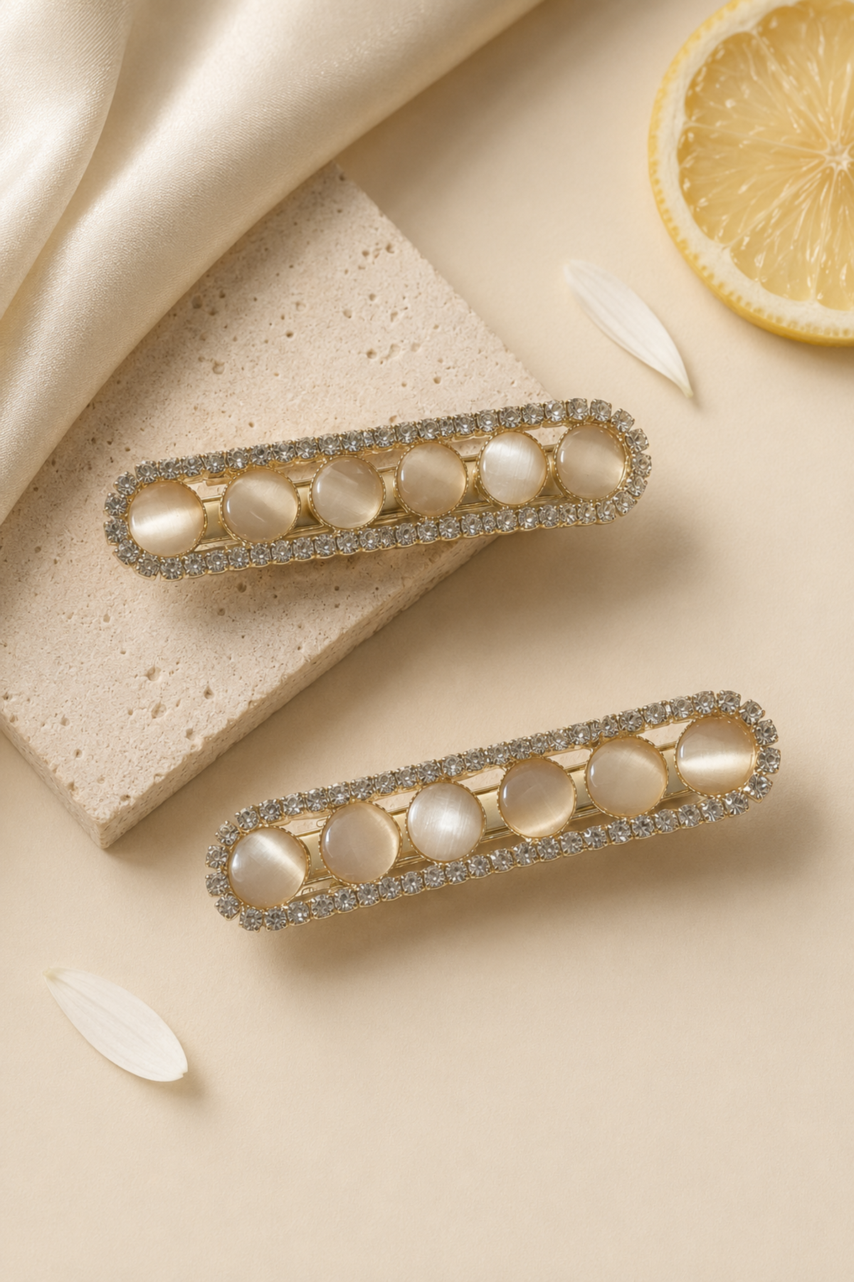 Two gold-toned barrette hair clips with oval pearl-like center stones and a border of clear rhinestones, arranged on a beige surface with satin fabric, a stone slab, and a lemon slice prop.