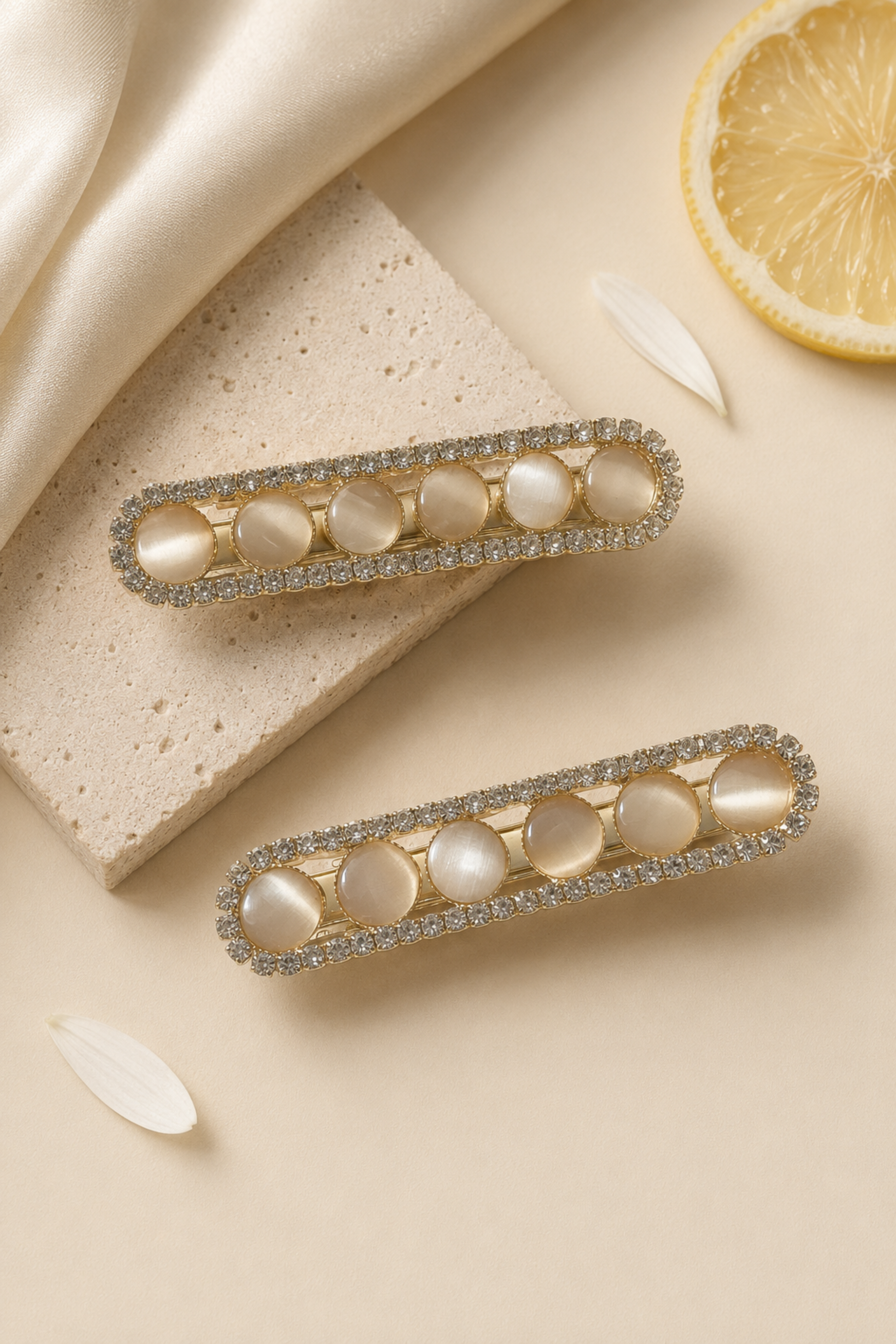 Two gold-toned barrette hair clips with oval pearl-like center stones and a border of clear rhinestones, arranged on a beige surface with satin fabric, a stone slab, and a lemon slice prop.