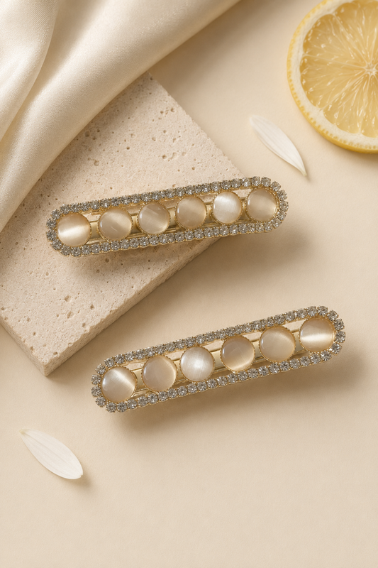 Two gold-toned barrette hair clips with oval pearl-like center stones and a border of clear rhinestones, arranged on a beige surface with satin fabric, a stone slab, and a lemon slice prop.