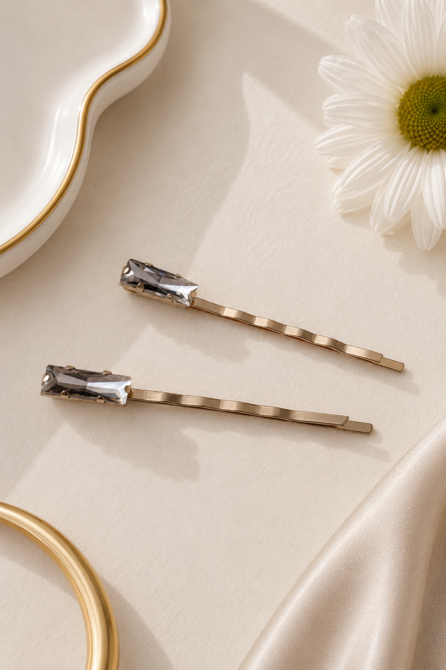 Two gold bobby pins with clear faceted crystal tips, arranged on a cream satin fabric next to a white daisy flower and gold-toned decorative props.