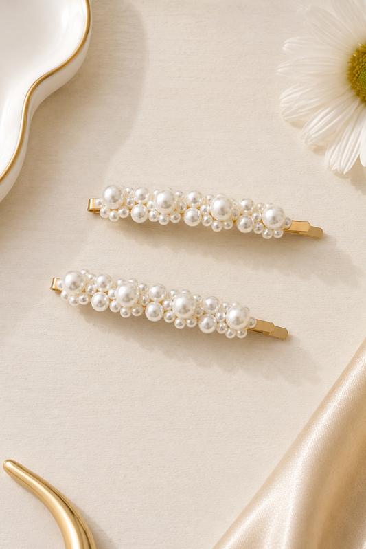 Two gold hair claw clips decorated with clusters of white faux pearls, arranged on a beige satin fabric background with a white daisy and gold dish as props.