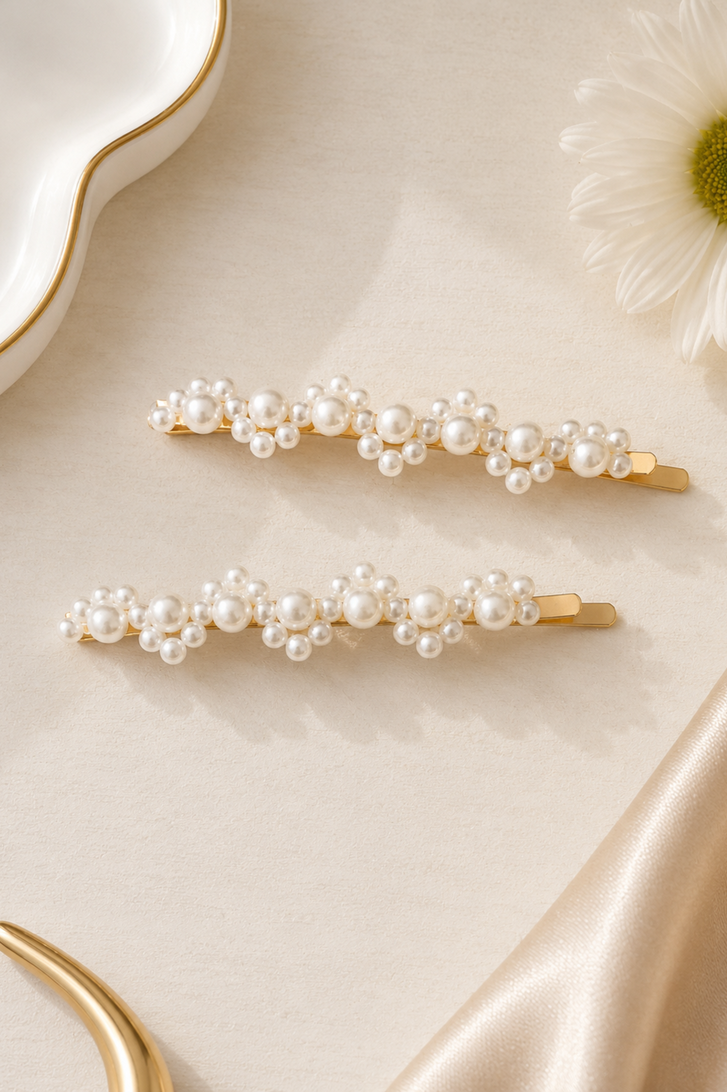 Two gold hair claw clips decorated with clusters of white faux pearls, arranged on a beige satin fabric background with a white daisy flower and gold dish accents.