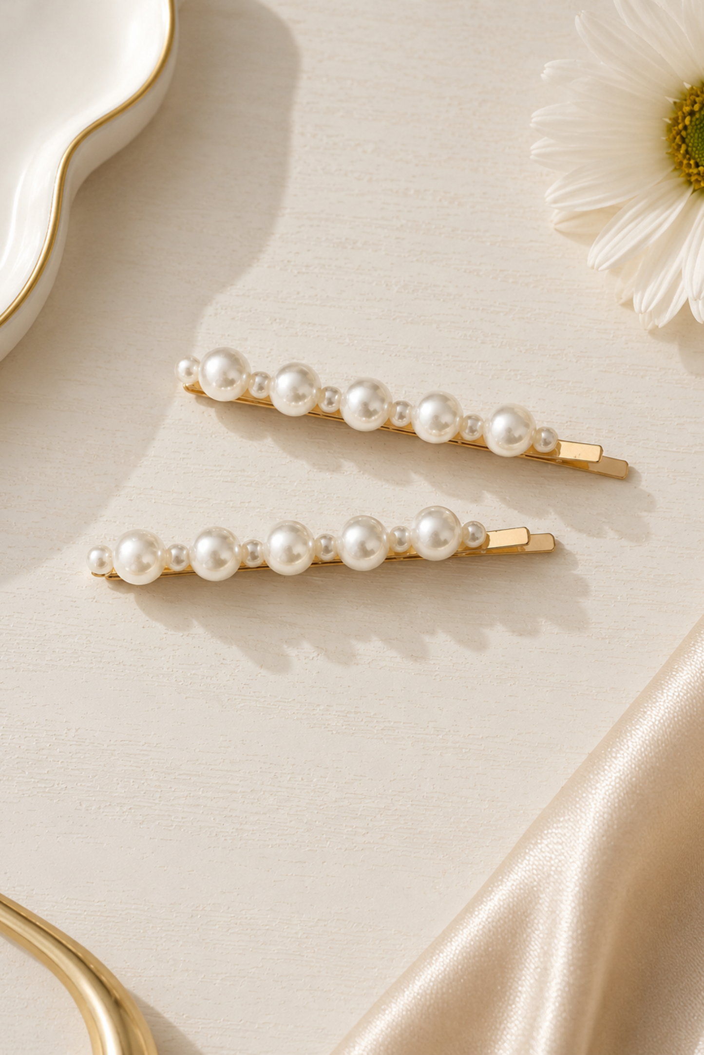 Two gold hair claw clips decorated with white faux pearls, arranged on a cream satin fabric with a white daisy flower and a white dish in the background.
