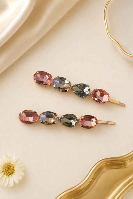 Two gold hair claw clips with faceted multicolor gemstones, styled on a cream satin background with a gold dish and a small daisy flower.