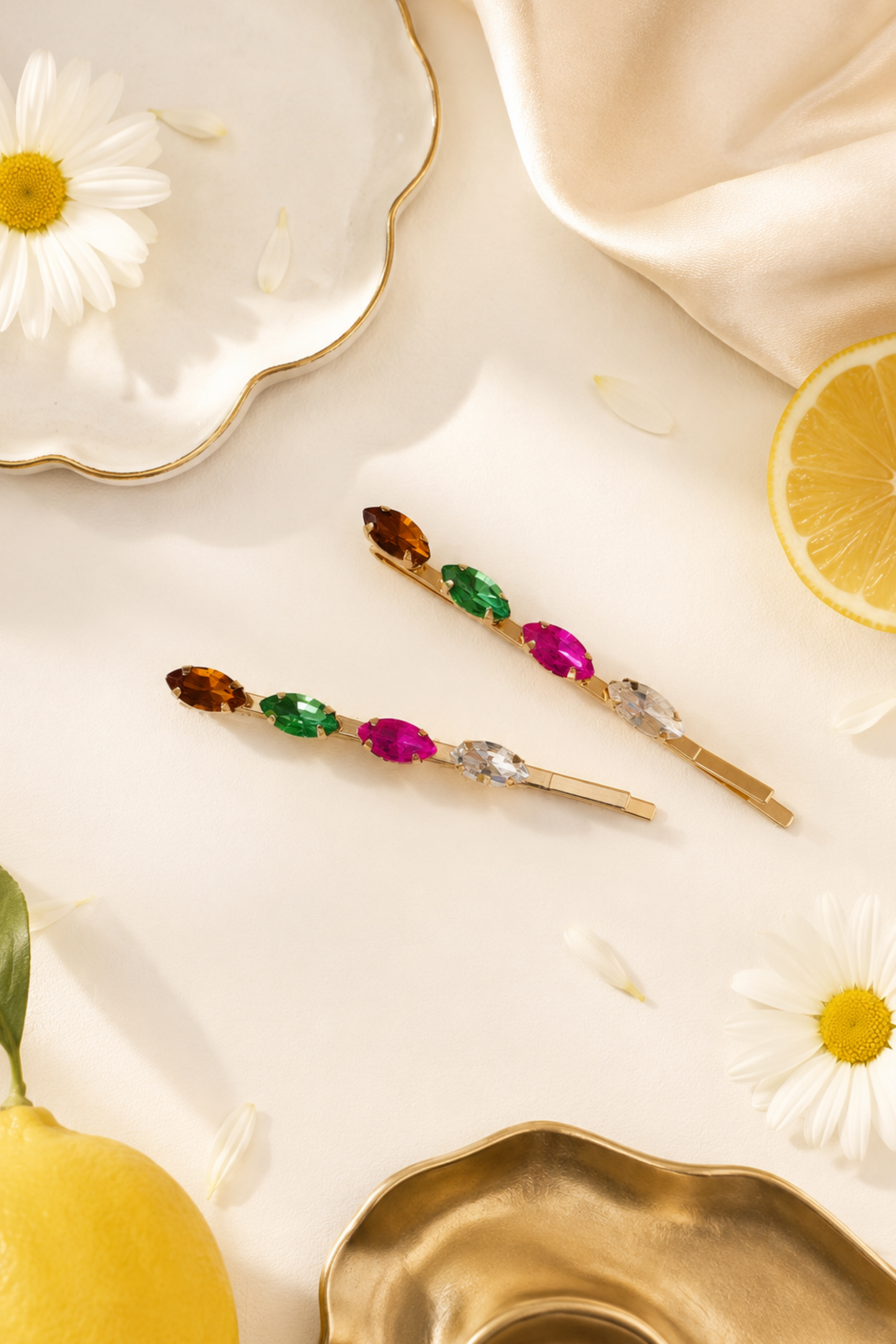 Two gold hair pins with colorful gemstone accents are arranged on a cream-colored surface with styled props including daisies, lemons, and gold-toned dishes.