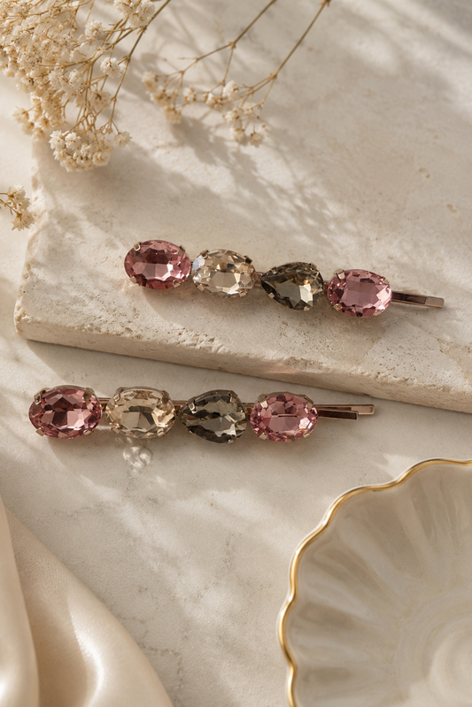 Two hair claw clips with faceted gemstone accents in pink, clear, and smoky gray, styled on a textured stone slab with dried flowers and cream satin.