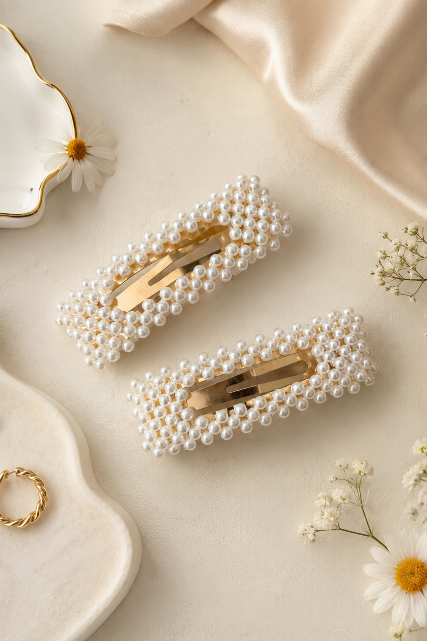 Two pearl-beaded hair clips with gold-tone metal clasps, arranged on a cream satin surface with decorative daisies and small jewelry pieces.
